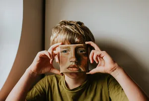 Un bambino tiene un piccolo dipinto a olio che lo raffigura sul viso Paralisi facciale: un bambino biondo tiene simbolicamente davanti al viso un ritratto dipinto
