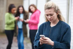 Testata_Intervista_Catarina_Katzer Cyberbullismo: una ragazza guarda tristemente il suo smartphone mentre i compagni di classe spettegolano su di lei sullo sfondo.