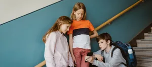p426m2525063 Drei Schulkinder stehen und sitzen auf einer Treppe und schauen auf ein Handy.