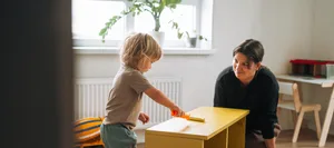 Une maman et son tout-petit peignent un meuble en bois en jaune à la maison, dans le cadre d’un projet de rénovation « DIY » avec les enfants Séparation : l'enfant joue avec un meuble en bois et la mère est assise à côté