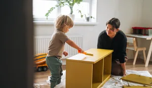Une maman et son tout-petit peignent un meuble en bois en jaune à la maison, dans le cadre d’un projet de rénovation « DIY » avec les enfants Séparation : l'enfant joue avec un meuble en bois et la mère est assise à côté