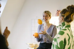 Une femme rit et jongle avec des oranges dans la cuisine Plan large montrant des amis debout dans la cuisine, tandis qu'une femme blonde, au centre, jongle avec des oranges en riant