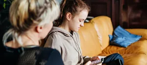 25-06-verhandlungen-swisscom-schweizer-elternmagazin-hg verhandeln: Mutter sitzt mit Teenager-Tochter, die aufs Handy schaut, auf dem Sofa