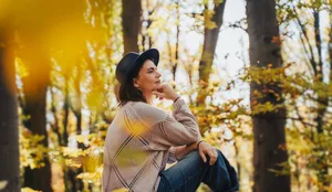 Smiling mid-adult perimenopausal Caucasian woman in a felt hat sitting on a log in the autumn forest. Frau in den Wechseljahren sitzt nachdenklich auf einem Baumstamm im Herbstwald