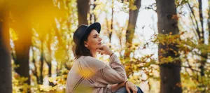 Smiling mid-adult perimenopausal Caucasian woman in a felt hat sitting on a log in the autumn forest. Frau in den Wechseljahren sitzt nachdenklich auf einem Baumstamm im Herbstwald