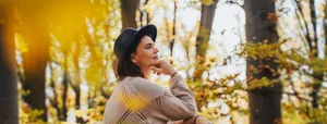 Smiling mid-adult perimenopausal Caucasian woman in a felt hat sitting on a log in the autumn forest. Frau in den Wechseljahren sitzt nachdenklich auf einem Baumstamm im Herbstwald