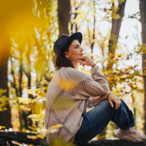 Smiling mid-adult perimenopausal Caucasian woman in a felt hat sitting on a log in the autumn forest. Frau in den Wechseljahren sitzt nachdenklich auf einem Baumstamm im Herbstwald