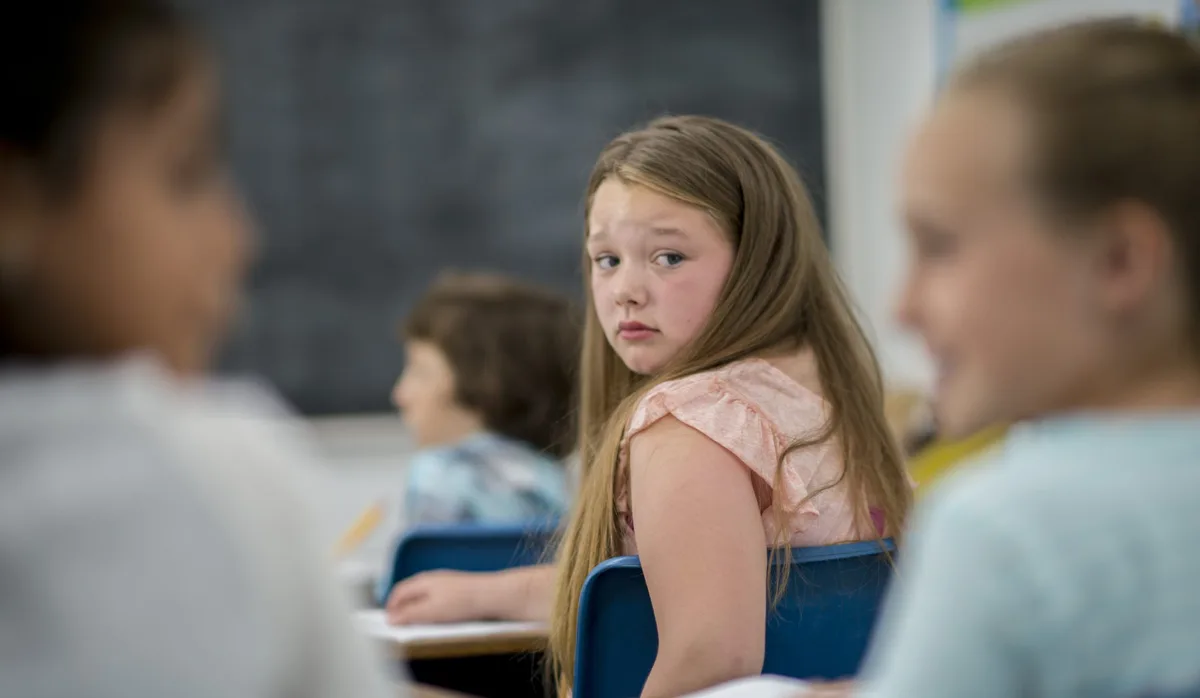 Mobbing in der Schule: Ein Mädchen schaut ängstlich nach hinten zu 2 tuschelnden Klassenkameradinnen Mobbing in der Schule: Ein Mädchen schaut ängstlich nach hinten zu 2 tuschelnden Klassenkameradinnen