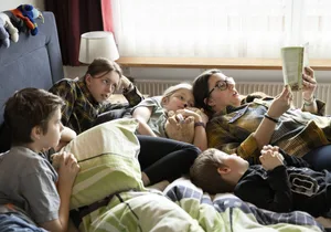 La famille Luigjes : Irina et Raymon avec Zimra (11 ans), Lias (10 ans), Moana (8 ans) et Niam (6 ans) ; moment en famille ; jeux en commun ; les enfants jouent ; sculpter ensemble, jouer au football, faire du trampoline Où va cette famille ? Une famille est allongée sur le lit, la mère fait la lecture à haute voix.