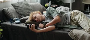 Teenage Caucasian Boy Lying on Bed Using Smartphone Suffering Gadget Addiction Social Media: Ein Junge liegt mit dem Handy auf dem Bett