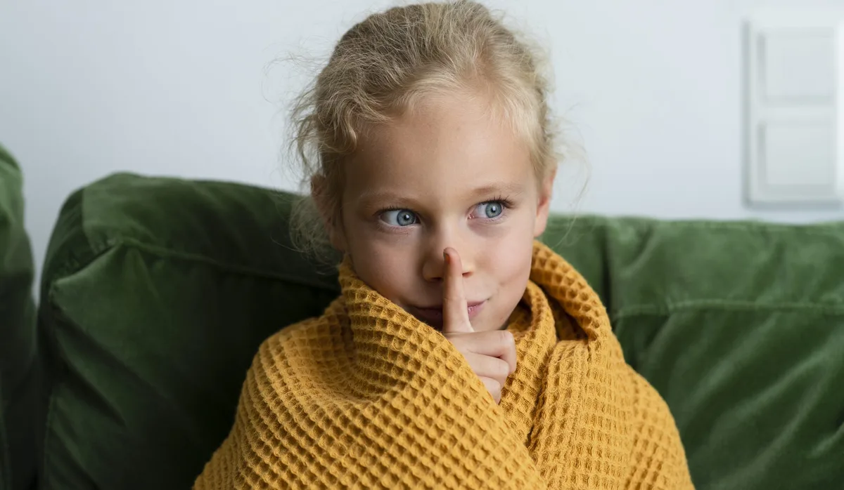 Contemplative girl wrapped in blanket making silence gesture at home Hochsensible Kinder reagieren oft empfindlicher auf Lärm