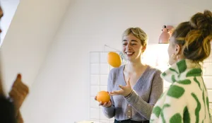 Woman laughs and juggles oranges in the kitchen Wide angle shot of friends standing in the kitchen while a blonde woman in the centre juggles oranges and laughs