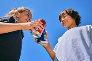 A boy and a girl drink carbonated drinks from cans, laugh and chat. Photo on the background of the sky. Zwei Kinder mit Energydrinks: Das macht eine Dose Red Bull mit dem Körper Ihres Kindes