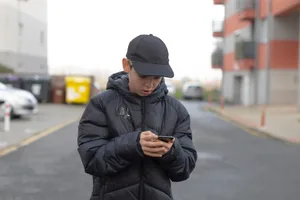A man walking along the street, engrossed in his smartphone, against a city backdrop under a grey sky Male teenager on his smartphone
